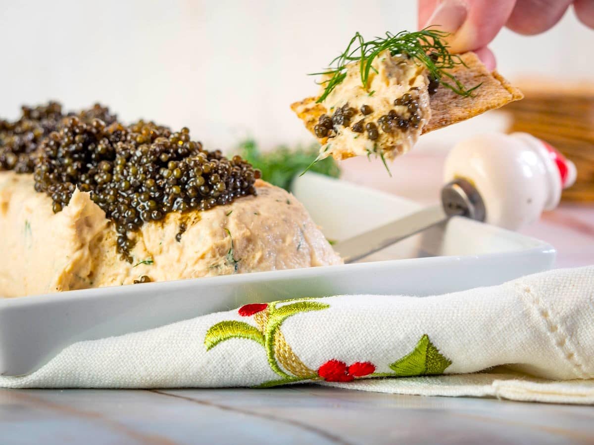 A hand spreads caviar-topped mousse onto a cracker over a rectangular white dish, with a napkin featuring red and green embroidery in the foreground.