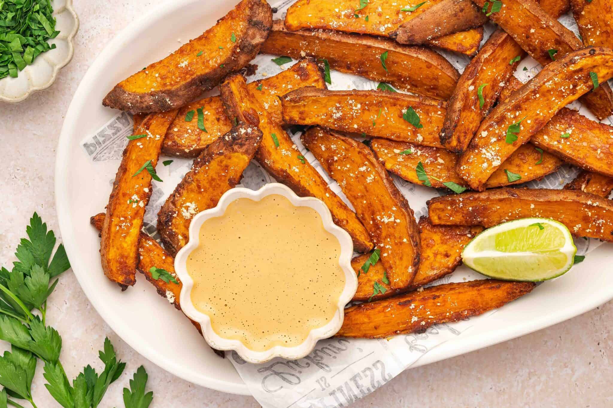 Plate of seasoned sweet potato fries garnished with parsley and salt, served with a creamy dipping sauce and a wedge of lime on the side.