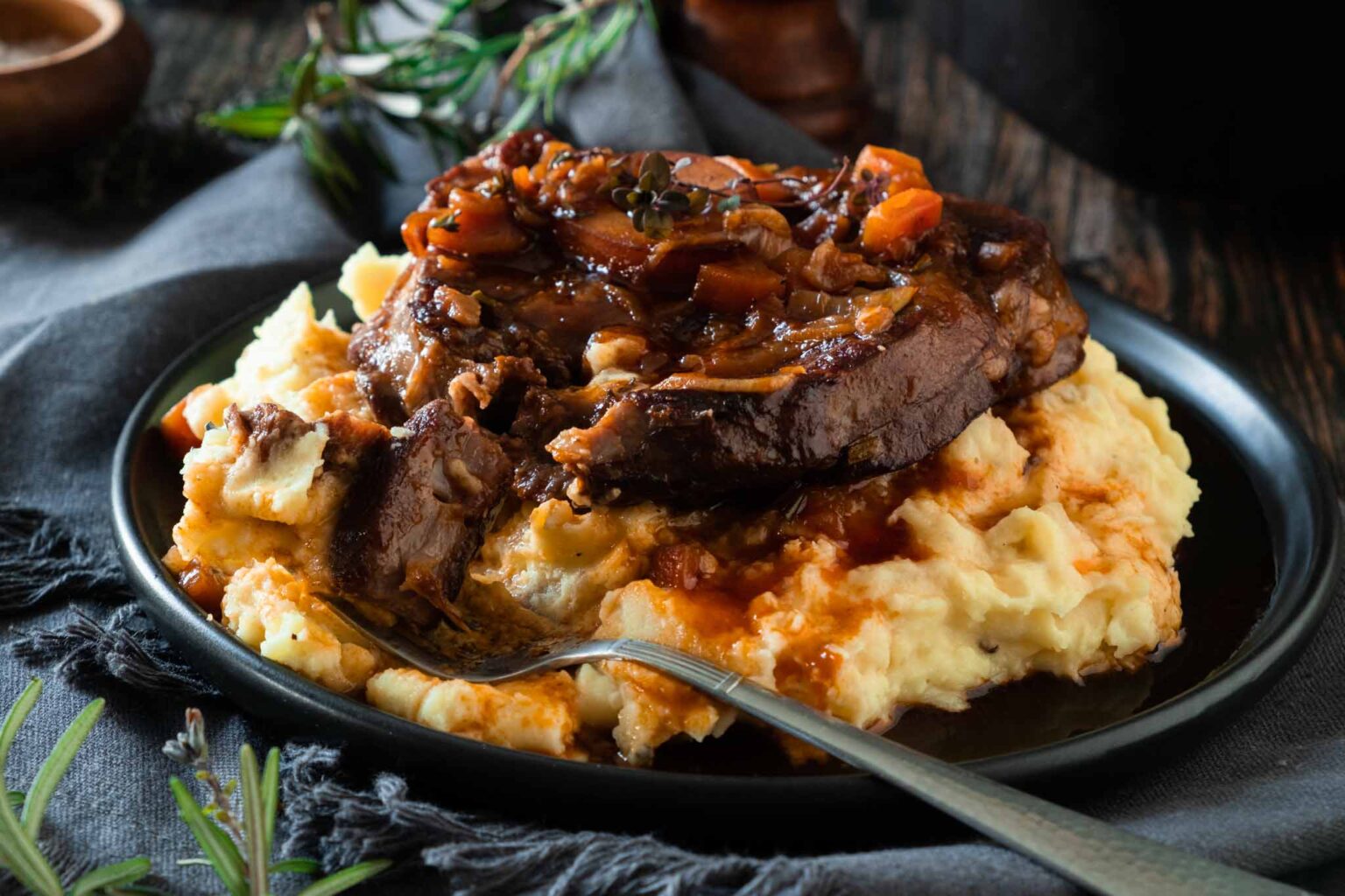 Braised beef with gravy and vegetables served over mashed potatoes on a black plate with a fork, garnished with herbs.