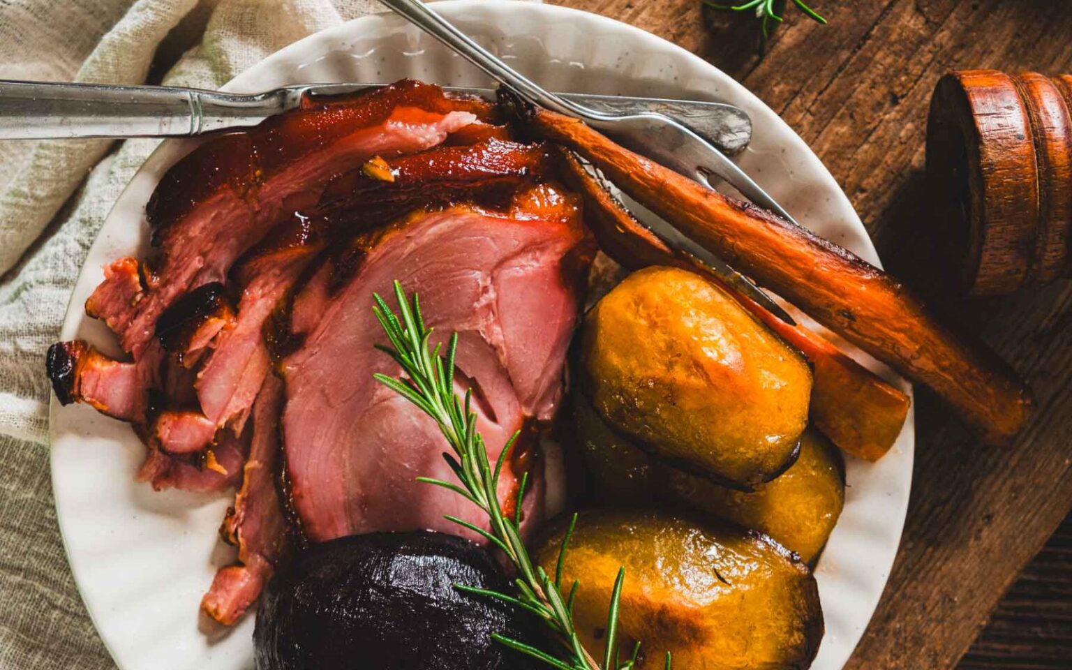 A plate with sliced glazed ham, roasted sweet potatoes, a sprig of rosemary, and cutlery on a wooden table.