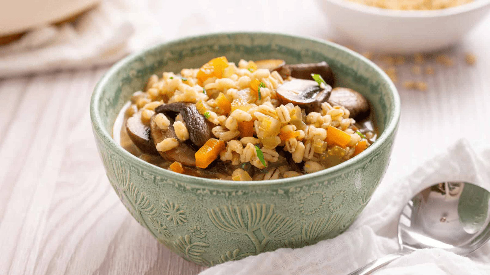 A green bowl filled with barley soup containing mushrooms, carrots, and herbs sits on a white surface next to a spoon.