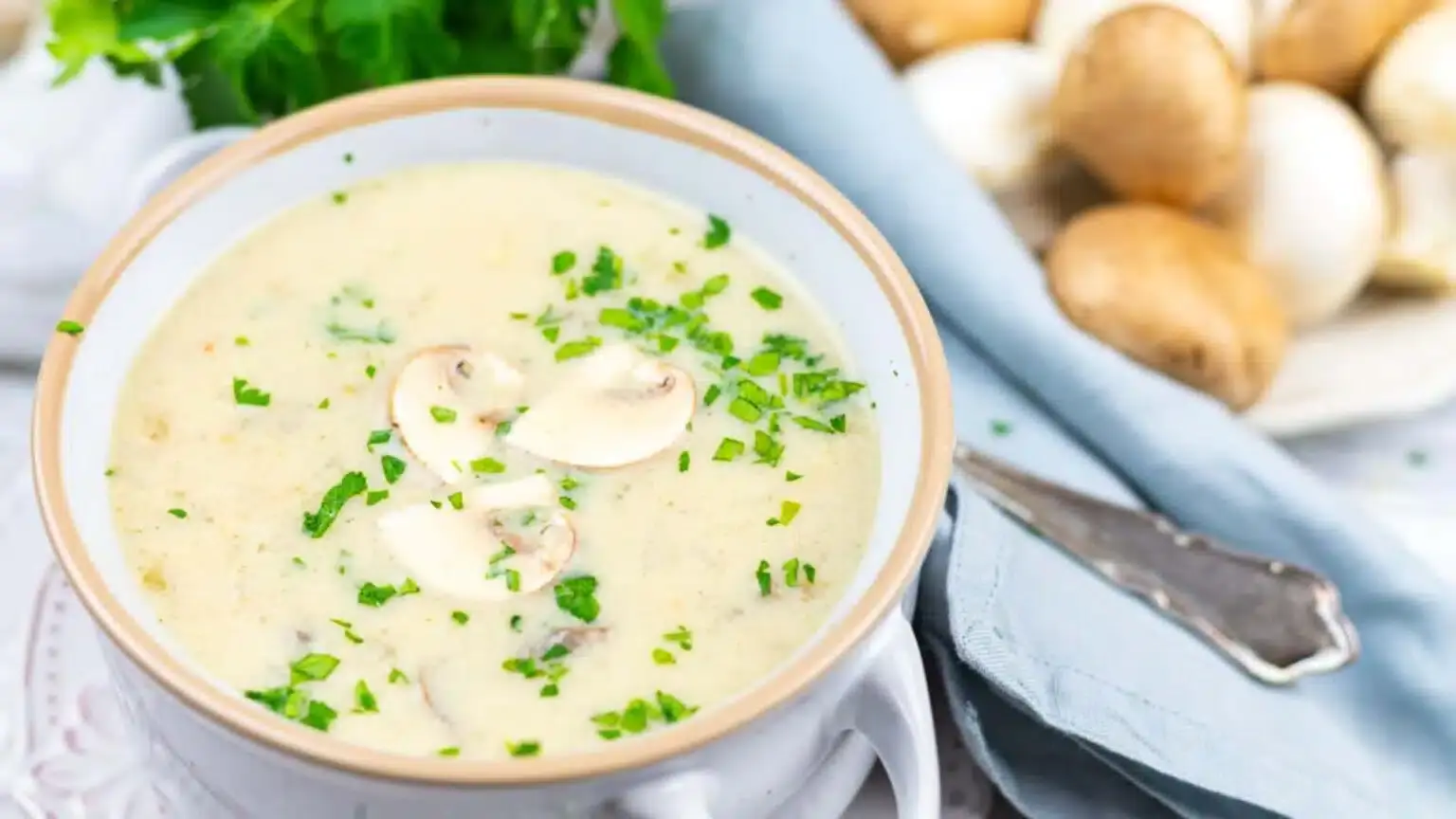 A bowl of creamy mushroom soup topped with fresh chopped herbs and sliced mushrooms, with whole mushrooms and parsley in the background.