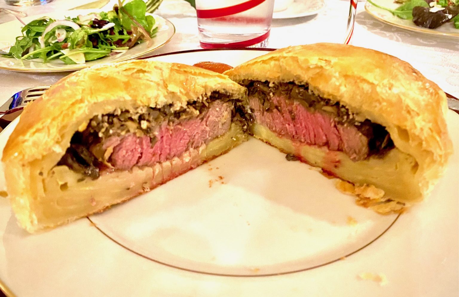A sliced Beef Wellington on a plate, showing layers of pastry, mushroom duxelles, and medium-rare beef, with a side salad in the background.