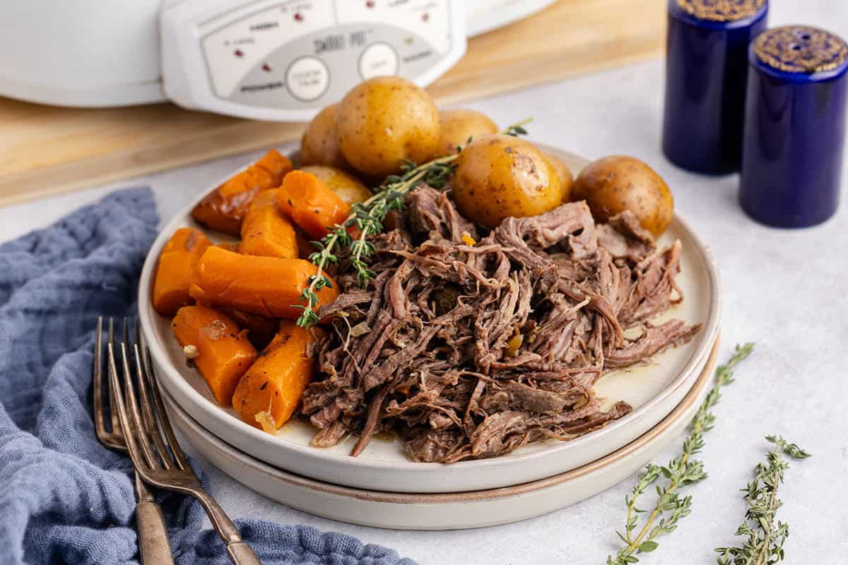 A plate of shredded pot roast with cooked carrots, whole potatoes, and a sprig of thyme, with a slow cooker and blue salt and pepper shakers in the background.