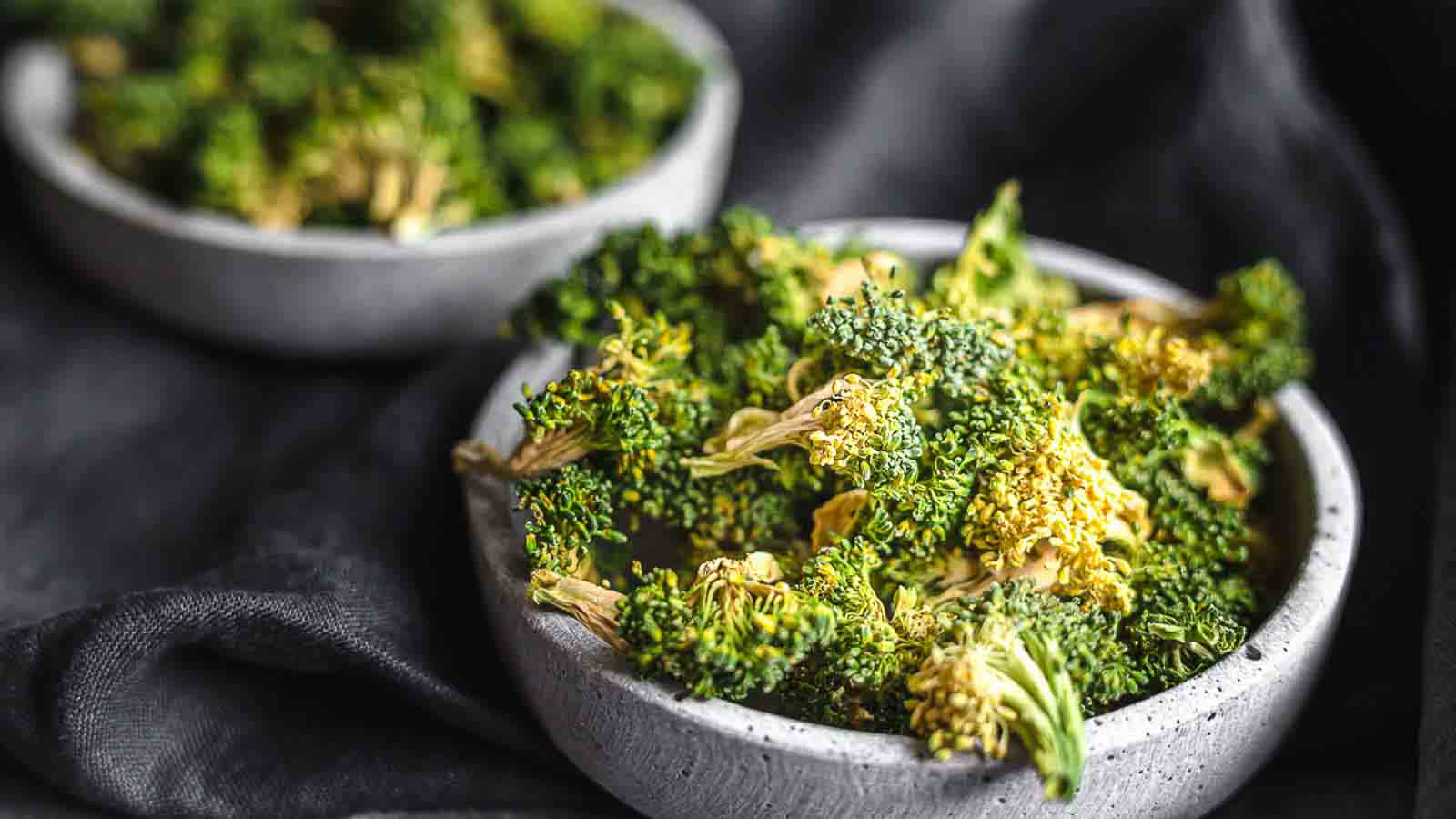 Two gray bowls filled with dried broccoli florets sit on a dark surface.