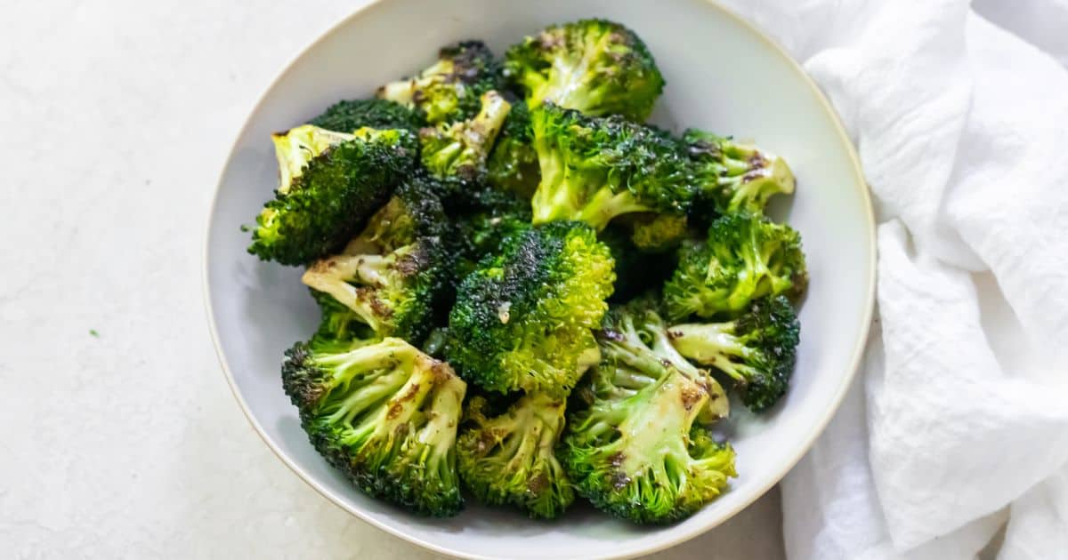 A white bowl filled with roasted broccoli florets sits on a light surface next to a white cloth.