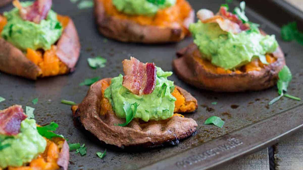 Baked sweet potato rounds topped with guacamole, bacon pieces, and chopped cilantro on a baking sheet.