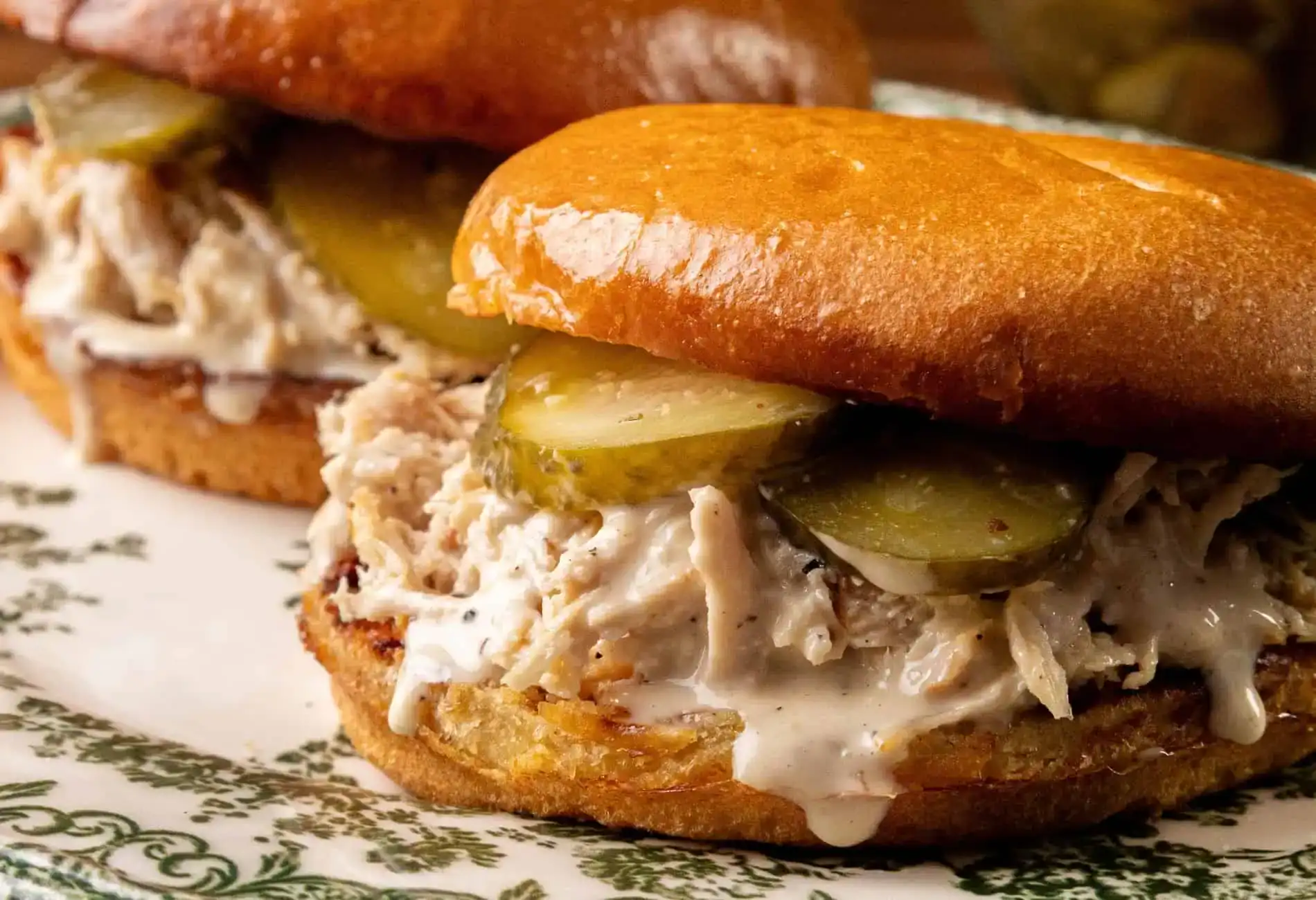 Two shredded chicken sandwiches with pickles and white sauce on toasted buns, served on a patterned plate.
