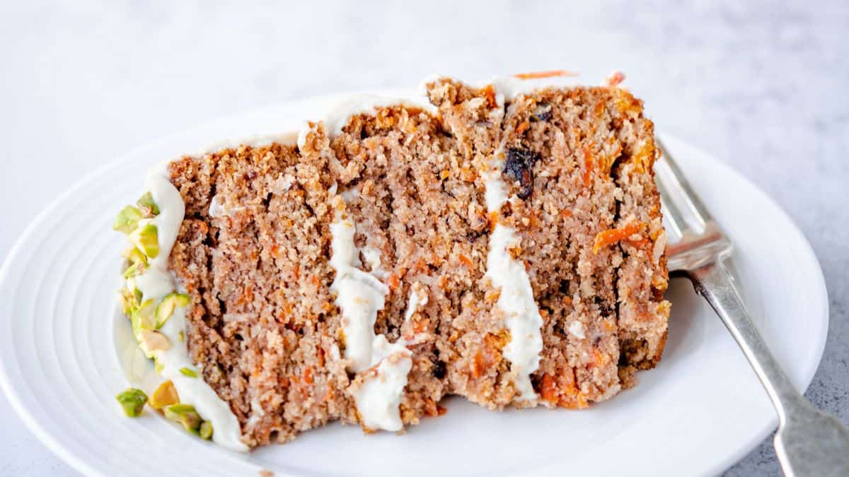 A slice of carrot cake with cream cheese frosting and chopped pistachios on the side, served on a white plate with a fork.