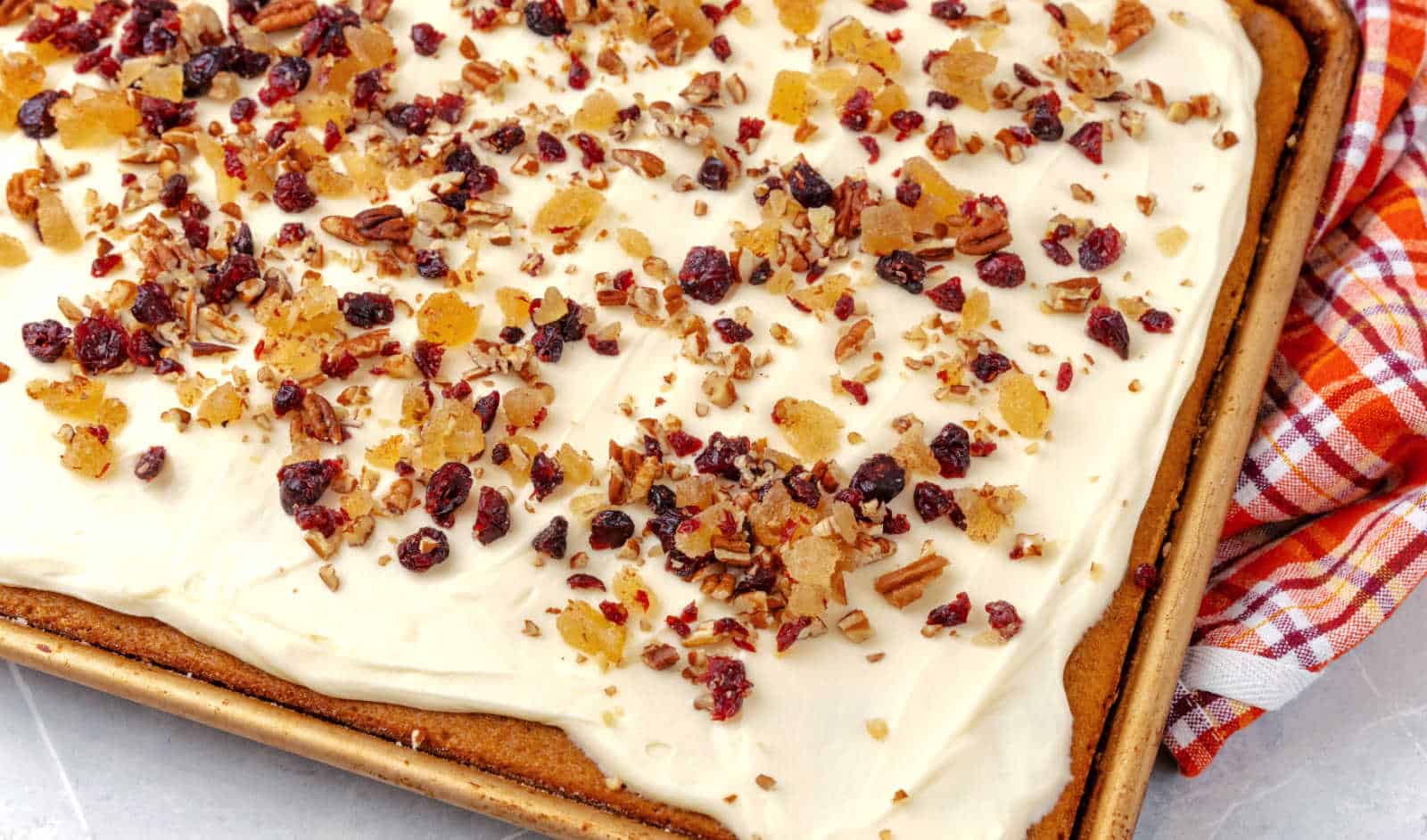 Sheet cake topped with white frosting, chopped pecans, dried cranberries, and candied ginger on a baking tray next to a red and white plaid cloth.