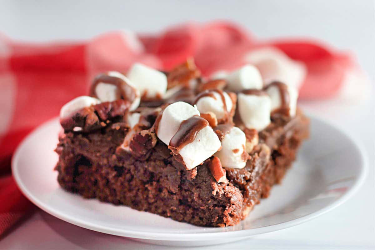 A slice of chocolate brownie topped with mini marshmallows, pecans, and a drizzle of chocolate sits on a white plate.