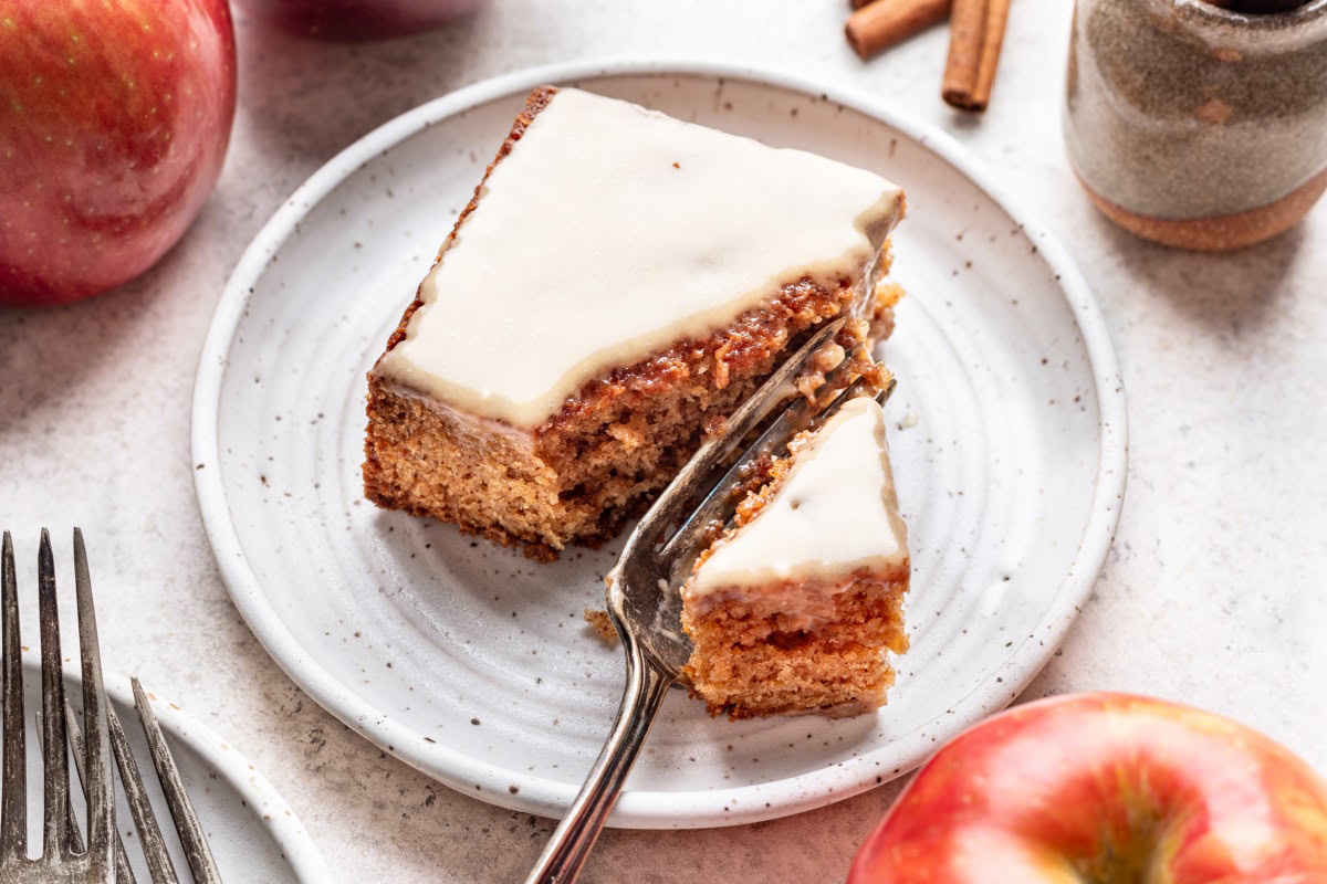 A slice of frosted apple cake on a white plate with a fork, surrounded by apples and cinnamon sticks.
