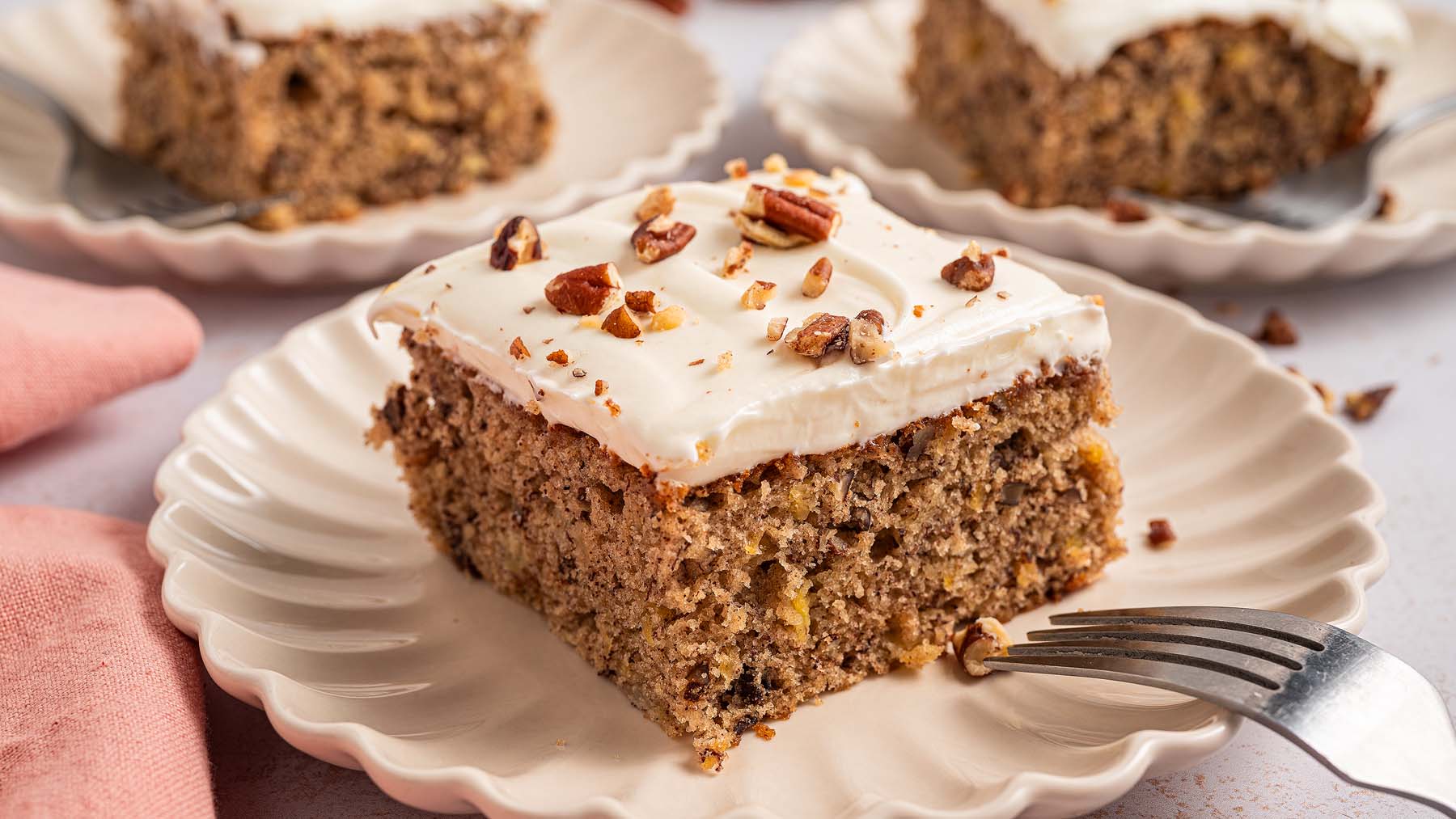 A slice of frosted cake topped with chopped nuts sits on a scalloped plate with a fork, with more slices visible in the background.