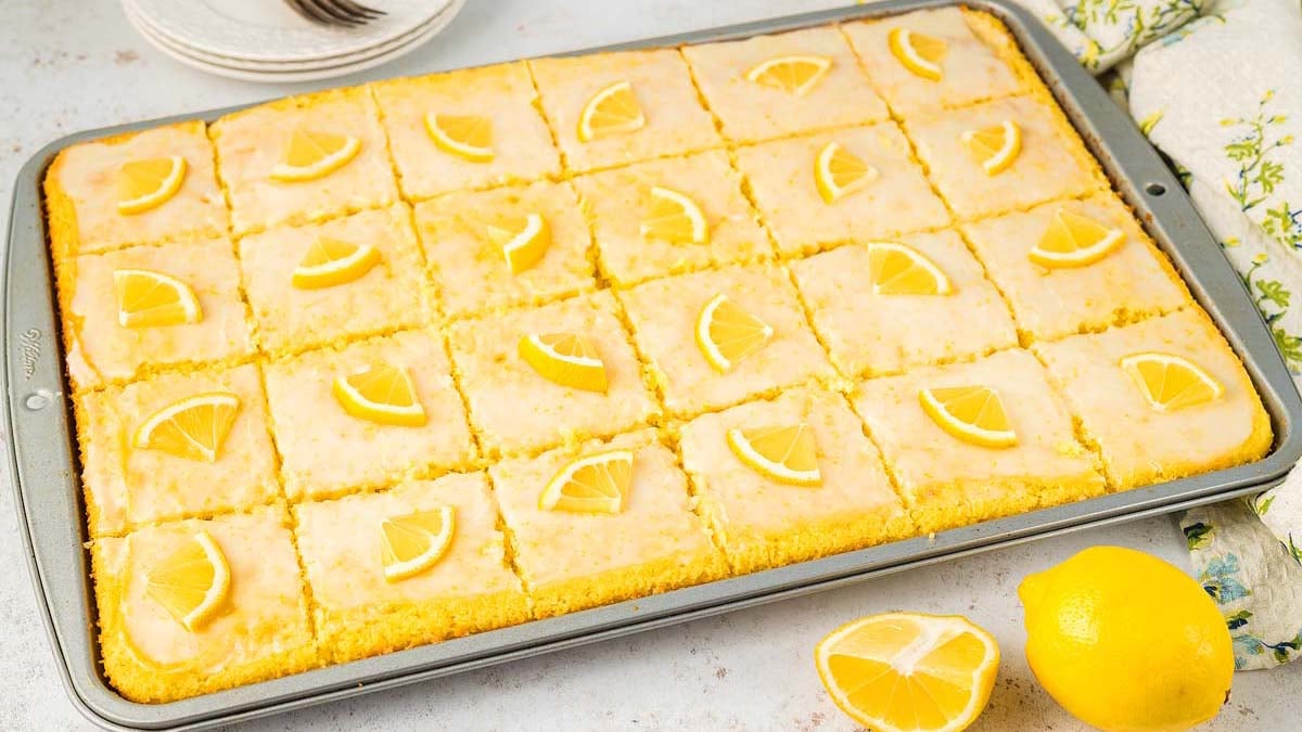 A baking tray with glazed lemon bars cut into squares, each topped with a small lemon wedge; a lemon and lemon wedges are next to the tray.