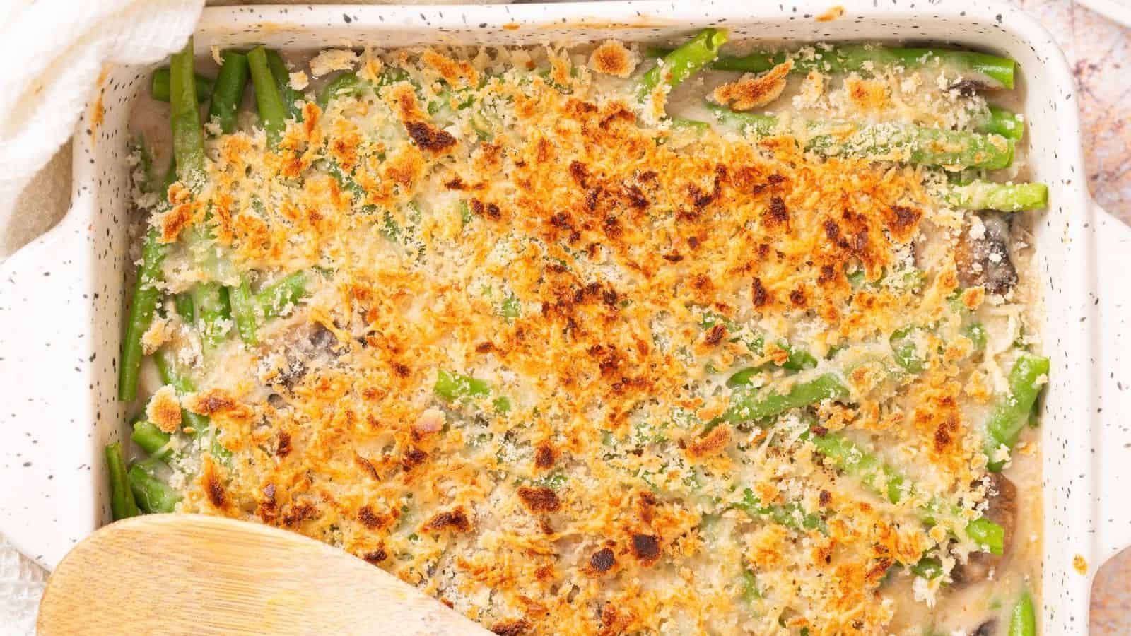 Baked green bean casserole topped with crispy breadcrumbs in a white ceramic dish, partially shown with a wooden spoon.