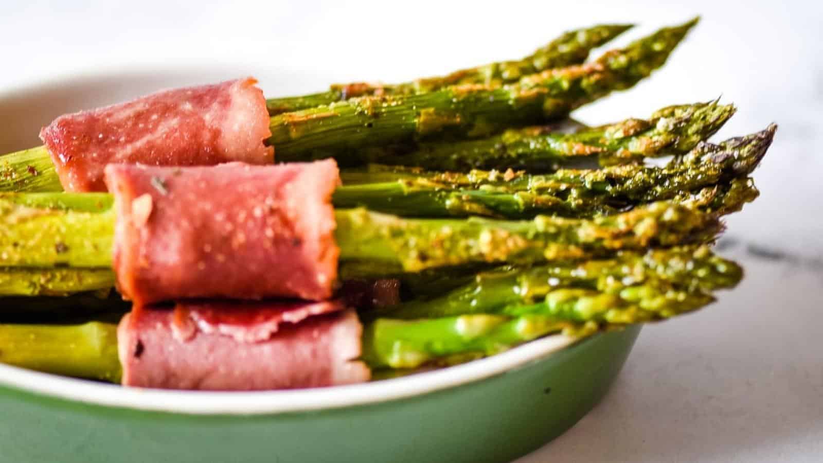 Several spears of cooked asparagus wrapped in slices of ham, served in a green dish.