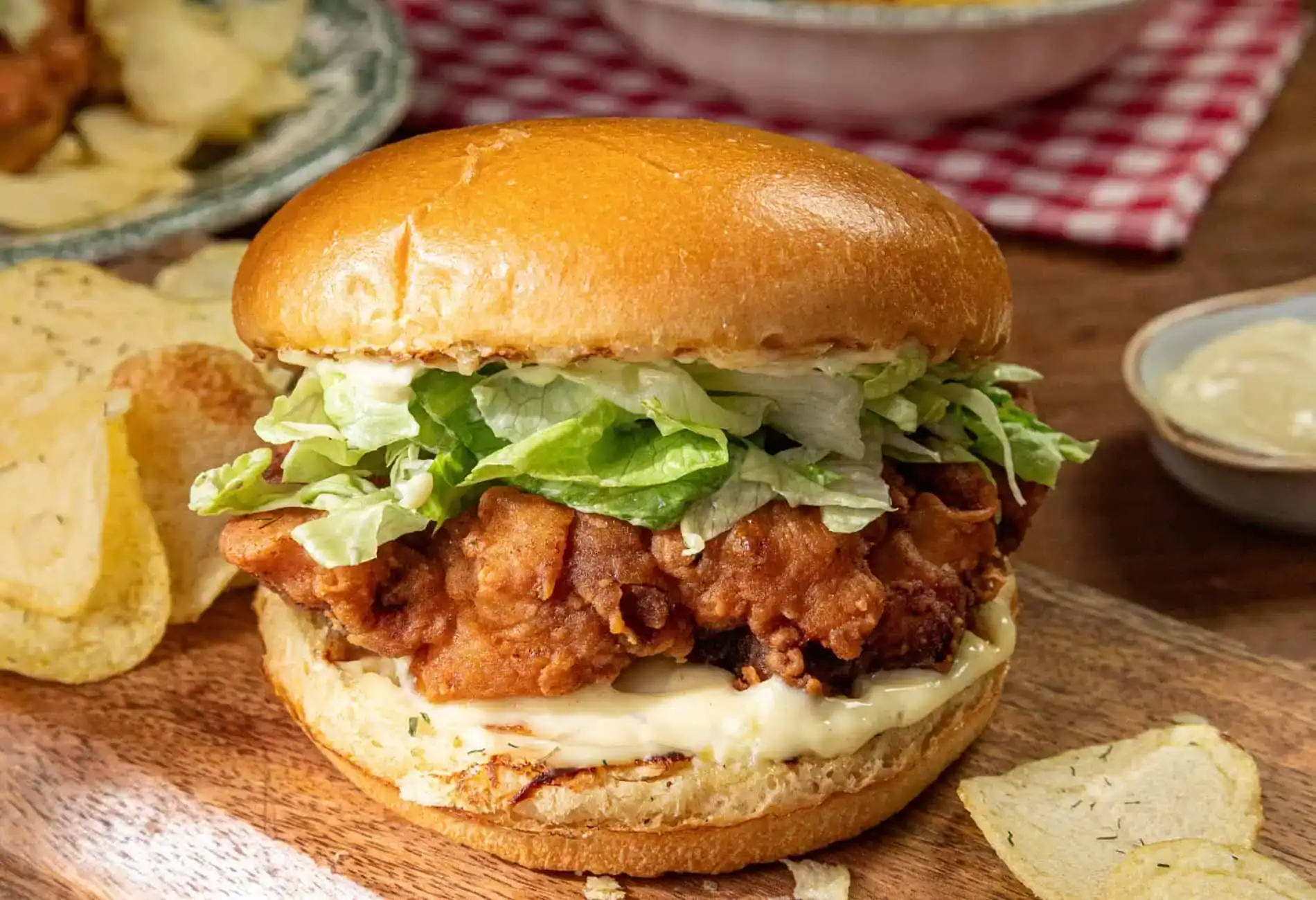 A fried chicken sandwich with lettuce and mayonnaise on a bun, served on a wooden board with potato chips on the side.