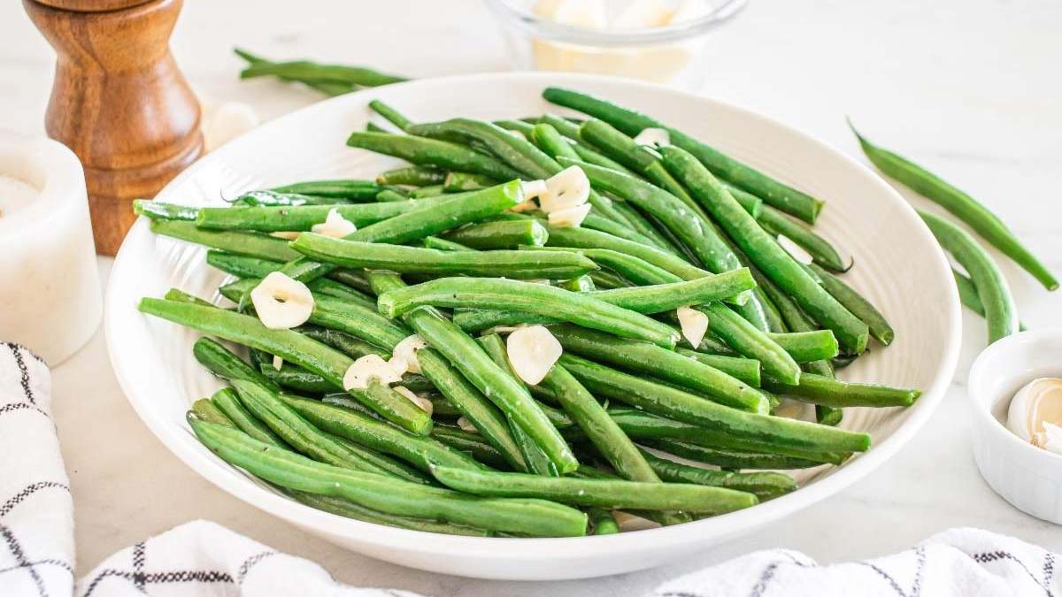 A white plate filled with cooked green beans garnished with sliced garlic, placed on a light surface next to a striped towel and a pepper grinder.