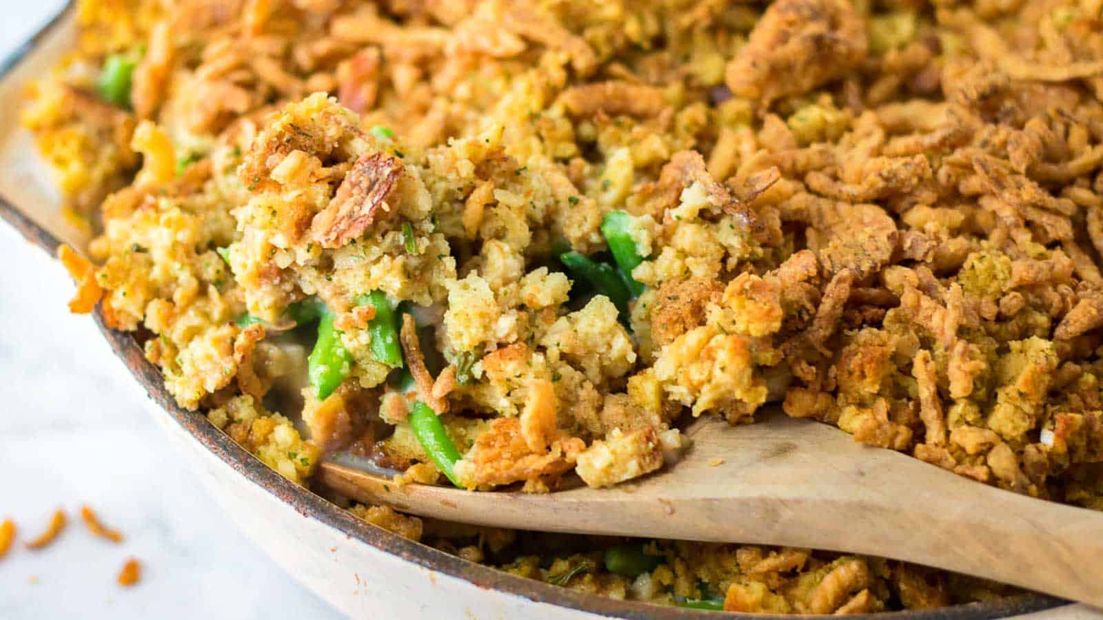 A close-up of a casserole dish with a wooden spoon scooping out a portion of green bean casserole topped with crispy breadcrumb and onion topping.