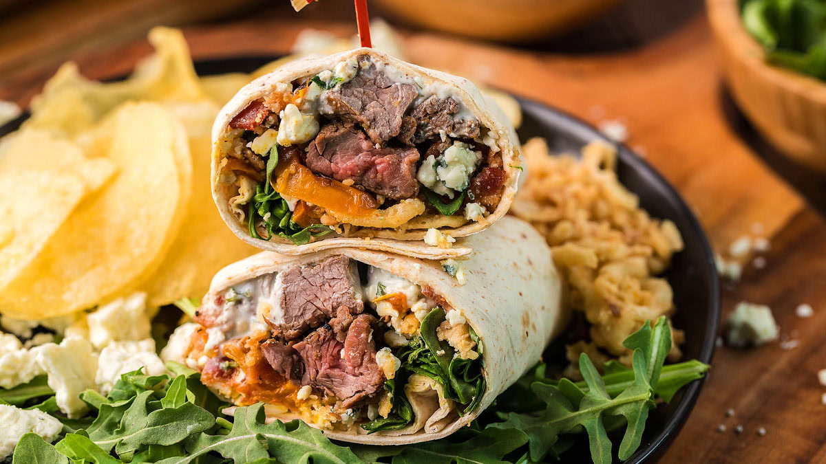 Two halves of a steak wrap filled with mixed greens, blue cheese, and tomatoes on a plate with potato chips, arugula, and crumbled cheese.