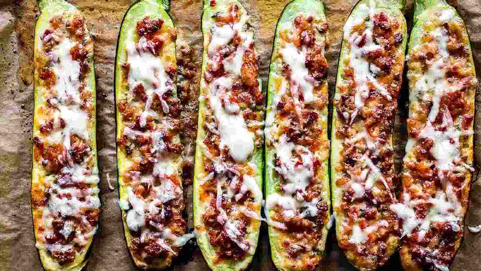 Six zucchini halves are arranged side by side, filled with a minced meat and tomato mixture, and topped with melted cheese on a baking sheet.