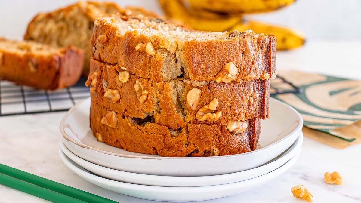 Three slices of banana nut bread stacked on a white plate, with more banana bread and ripe bananas visible in the background.