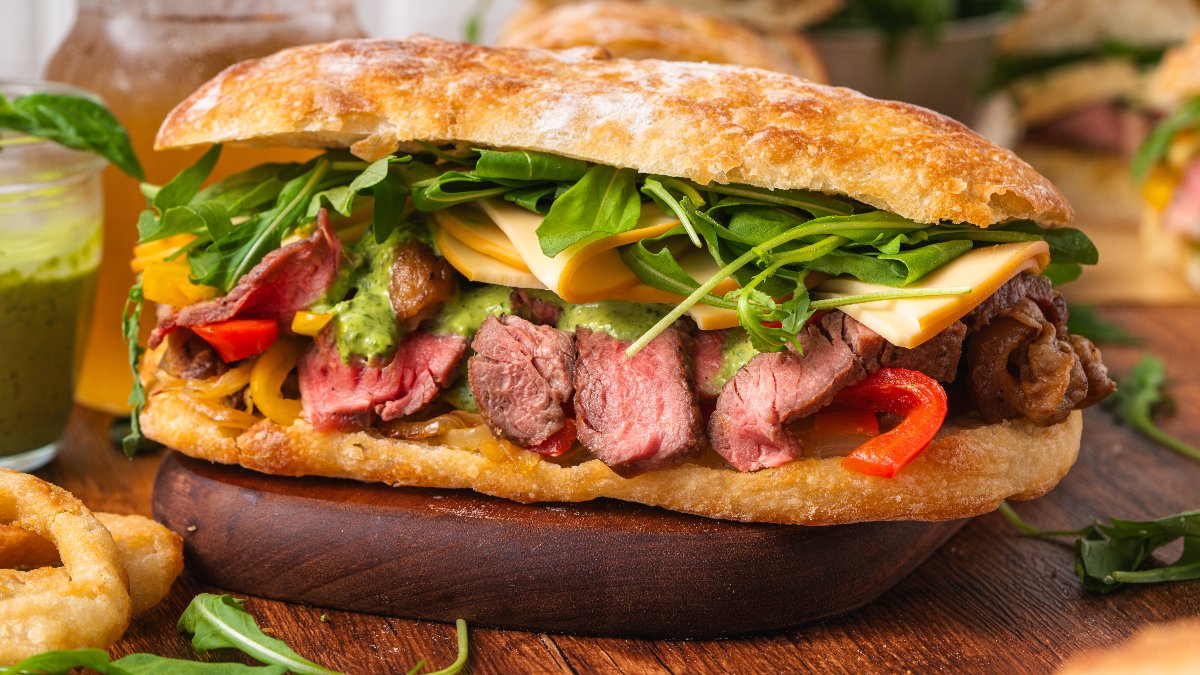 A steak sandwich with sliced beef, cheese, arugula, grilled peppers, and sauce on ciabatta bread, served on a wooden board.