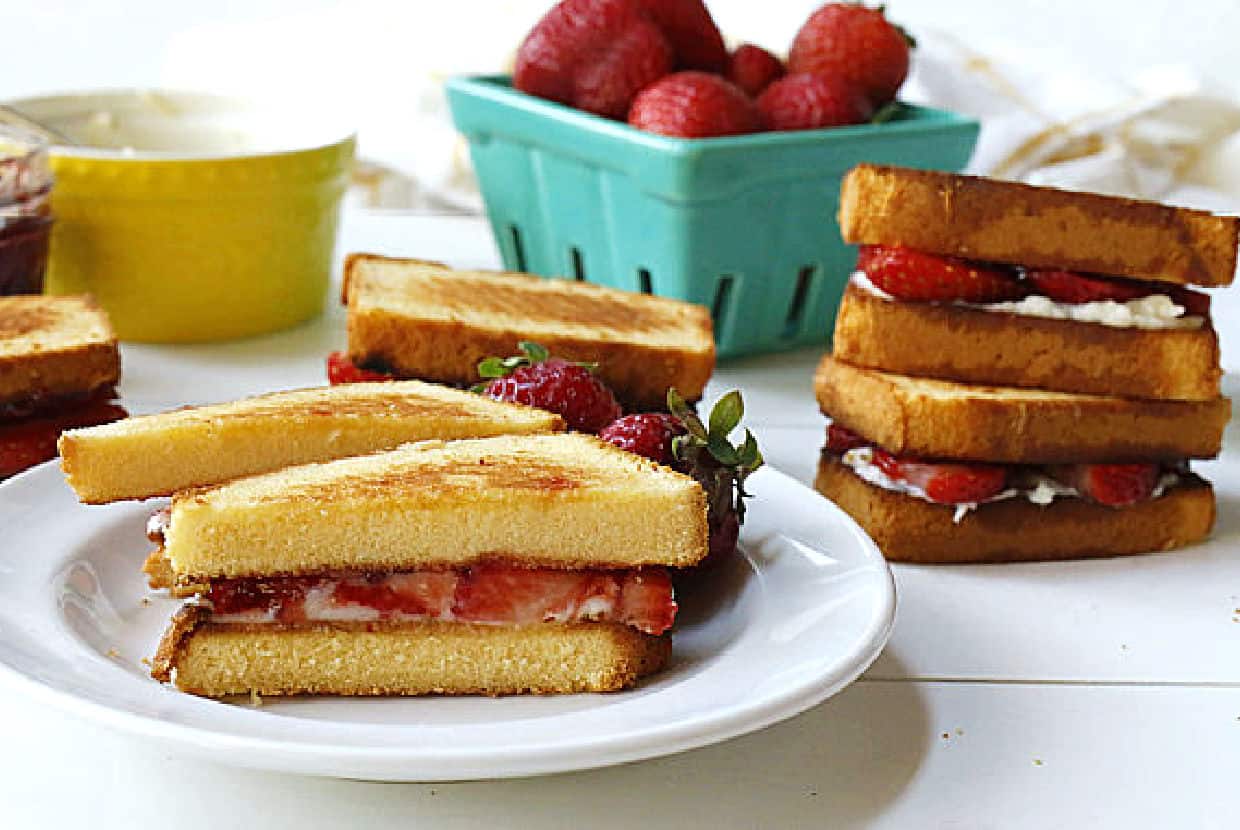 A plate with a halved strawberry sandwich, fresh strawberries, and stacked toast sandwiches with strawberry filling; a container of strawberries is in the background.