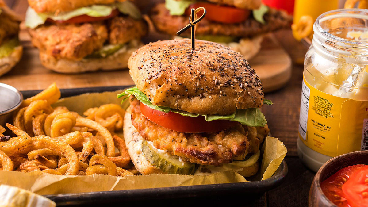 A breaded chicken sandwich with lettuce, tomato, and pickles on a seeded bun is served with curly fries on a tray. A jar of mustard and other sandwiches are in the background.