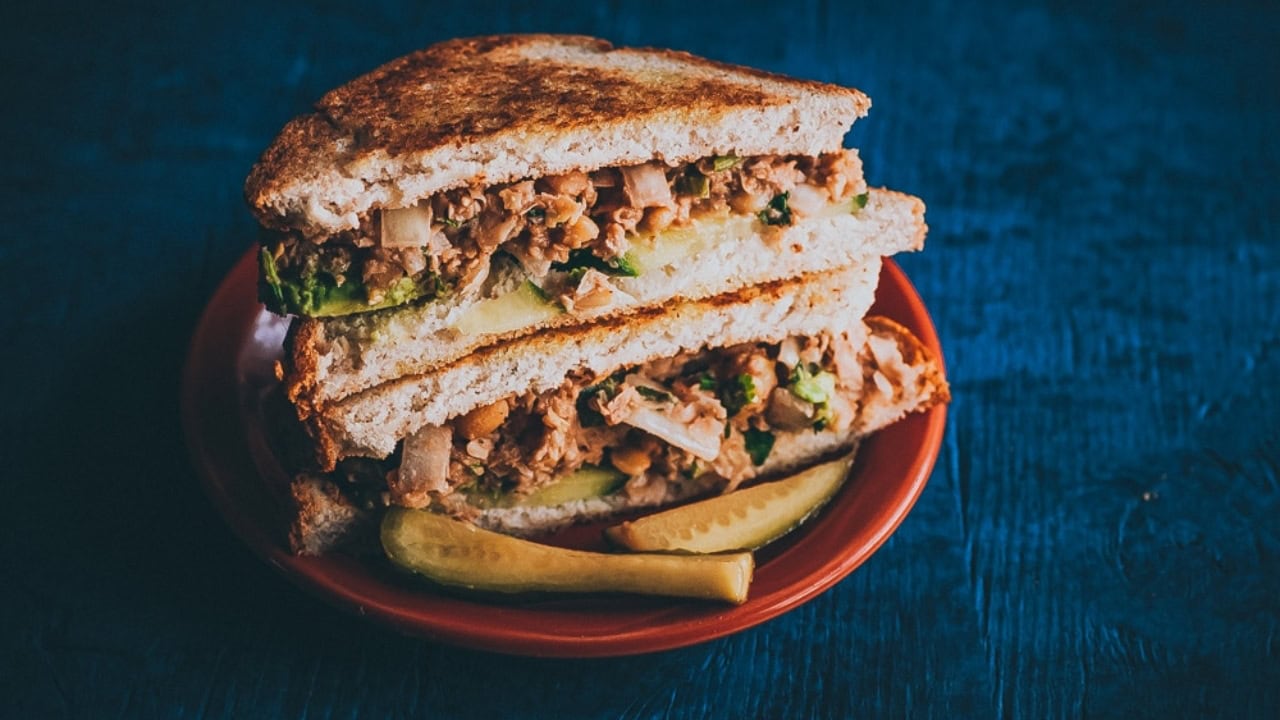 A tuna salad sandwich cut in half is stacked on a red plate with two pickle spears, set on a dark blue surface.