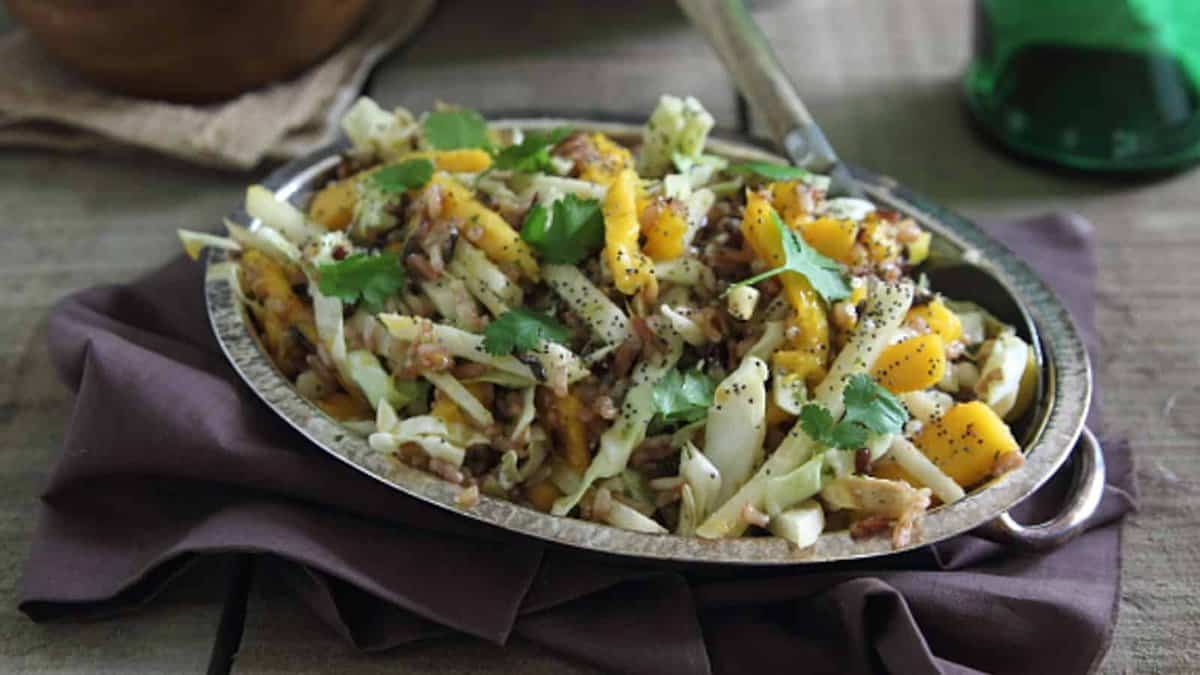 A metal dish filled with cabbage slaw mixed with mangoes, walnuts, poppy seeds, and garnished with fresh cilantro sits on a brown napkin.