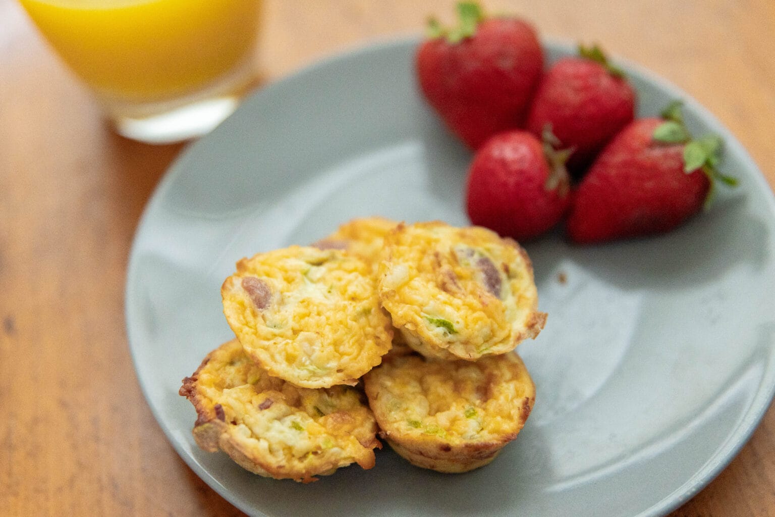 A gray plate with four egg muffins and three strawberries next to a glass of orange juice on a wooden table.