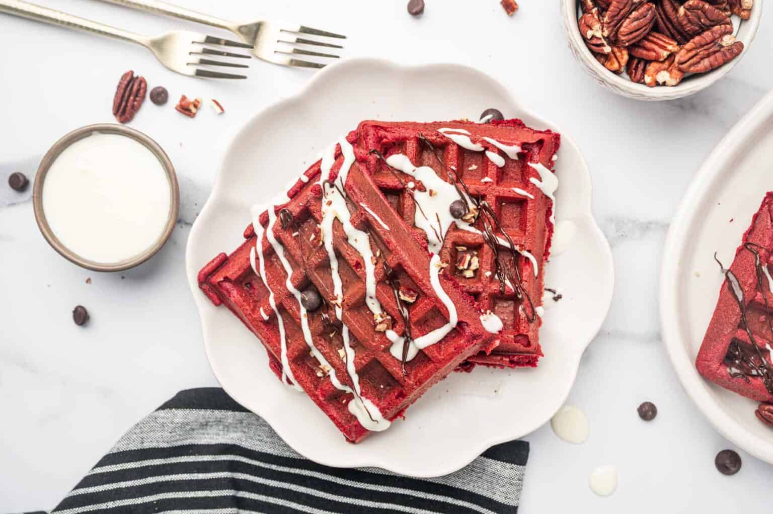 Two red velvet waffles topped with chocolate chips, pecans, and white icing on a white plate, with a glass of milk and a bowl of pecans nearby.