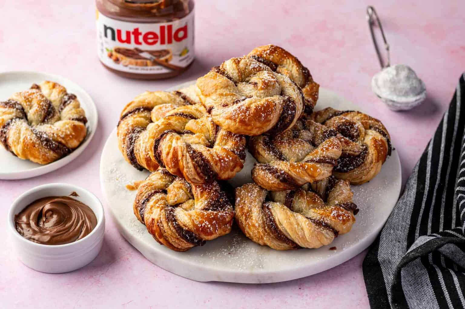 A plate of twisted pastries dusted with powdered sugar, with a bowl of Nutella, a jar of Nutella, and a sifter on a pink surface.
