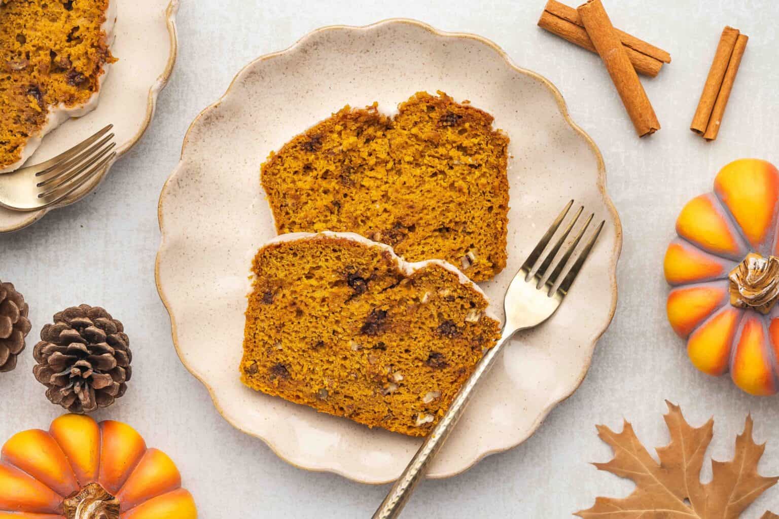 Two slices of pumpkin bread on a plate with a fork, surrounded by mini pumpkins, pinecones, cinnamon sticks, and a dried leaf.
