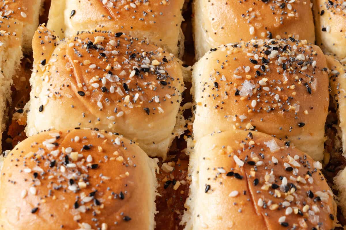Close-up of several golden brown slider buns topped with everything bagel seasoning, arranged closely together.