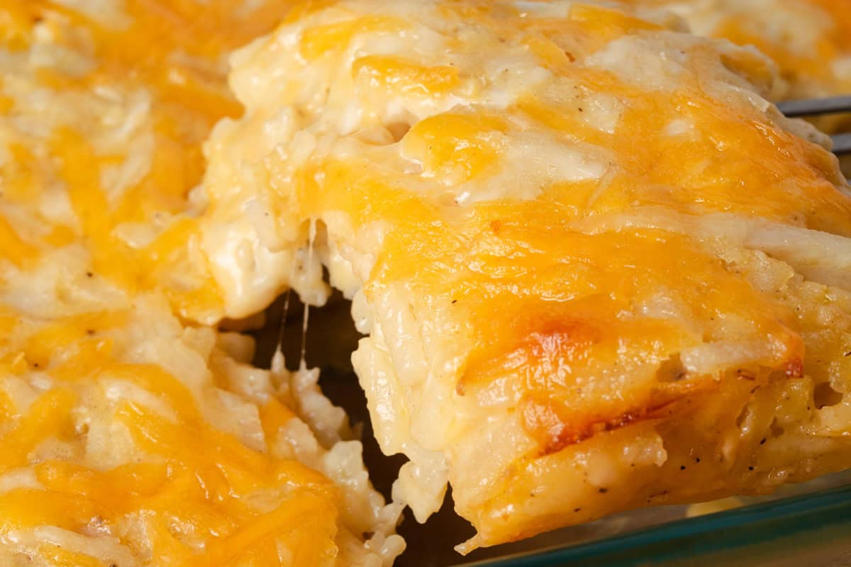 A close-up of a cheesy hash brown casserole, with a serving being lifted to show melted cheese and grated potatoes.