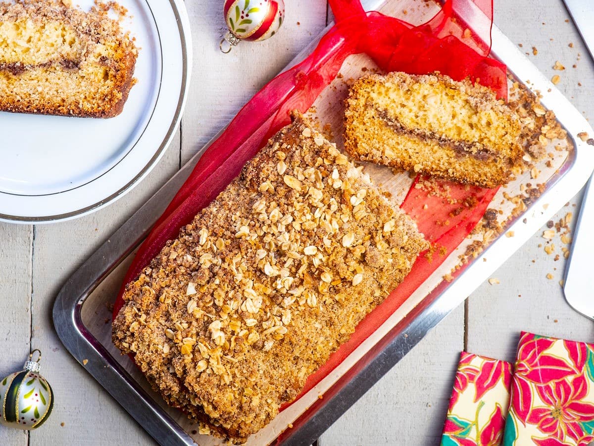 A loaf of crumb-topped coffee cake with a cinnamon layer is sliced on a tray with a red cloth, next to a plate and holiday ornaments.