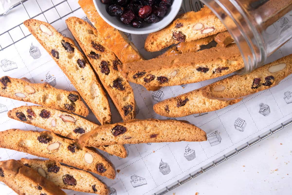 Sliced biscotti with almonds and dried cranberries are arranged on a wire rack, with a tipped jar and a small bowl of dried cranberries beside them.
