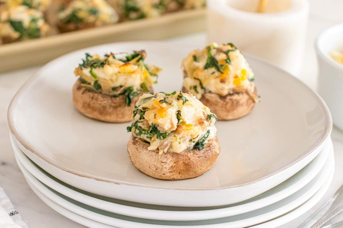 Three stuffed mushrooms with a creamy filling and herbs are arranged on a white plate, with more stuffed mushrooms blurred in the background.