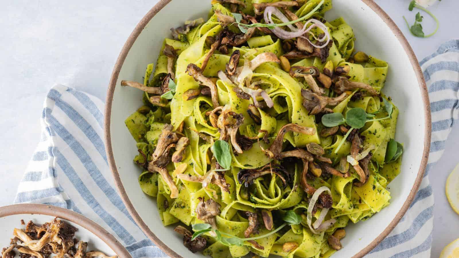 A bowl of wide ribbon pasta with pesto sauce, topped with sautรฉed mushrooms, thinly sliced onions, and fresh herbs, placed on a striped cloth.