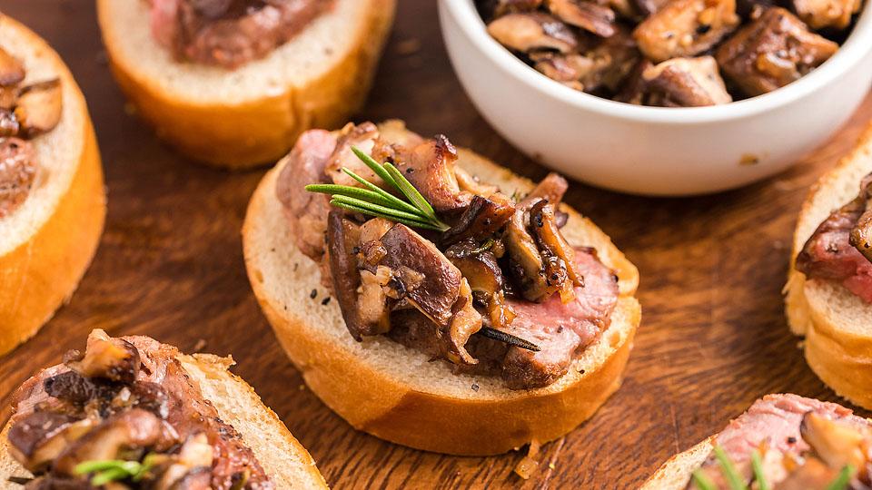 Slices of baguette topped with steak, sautรฉed mushrooms, and a sprig of rosemary are arranged on a wooden surface; a bowl of mushrooms is visible in the background.
