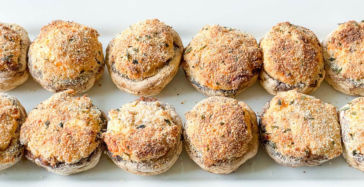 Twelve stuffed mushrooms topped with golden breadcrumbs are arranged in neat rows on a white rectangular platter.