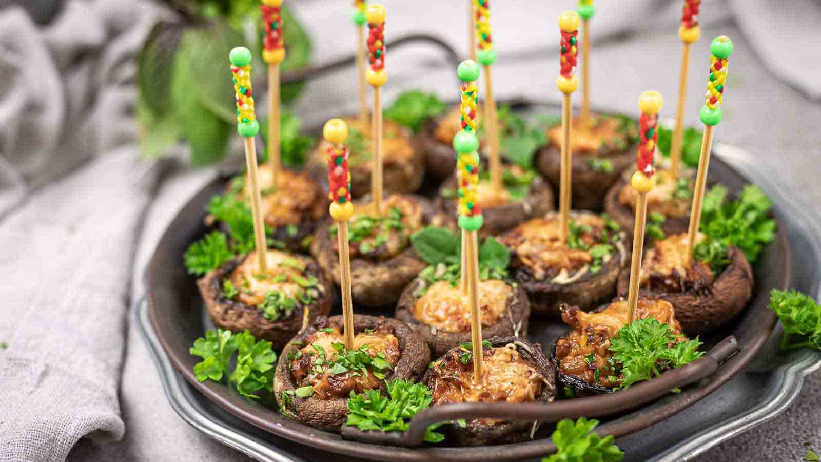 Stuffed mushrooms topped with herbs and cheese, each held with a colorful decorative toothpick, arranged on a dark plate with parsley garnish.