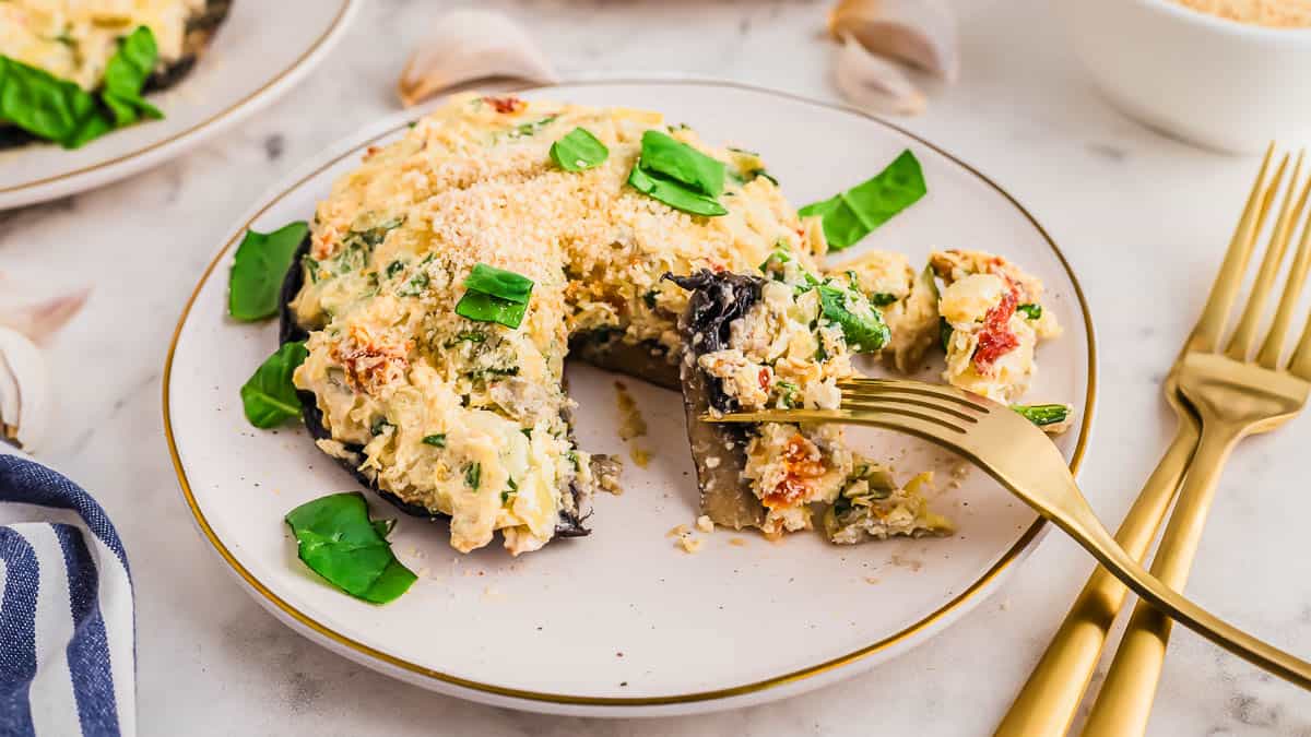 A stuffed portobello mushroom topped with a creamy mixture and fresh spinach, served on a white plate with gold utensils.