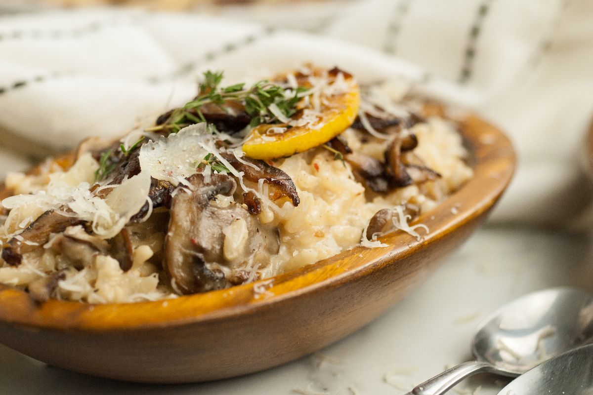 A wooden bowl filled with creamy mushroom risotto, garnished with a lemon slice, fresh herbs, and grated cheese.