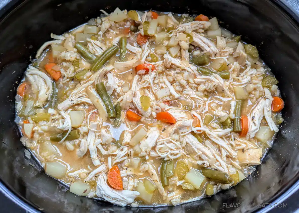 Shredded chicken, green beans, carrots, potatoes, and broth cooking in a slow cooker.