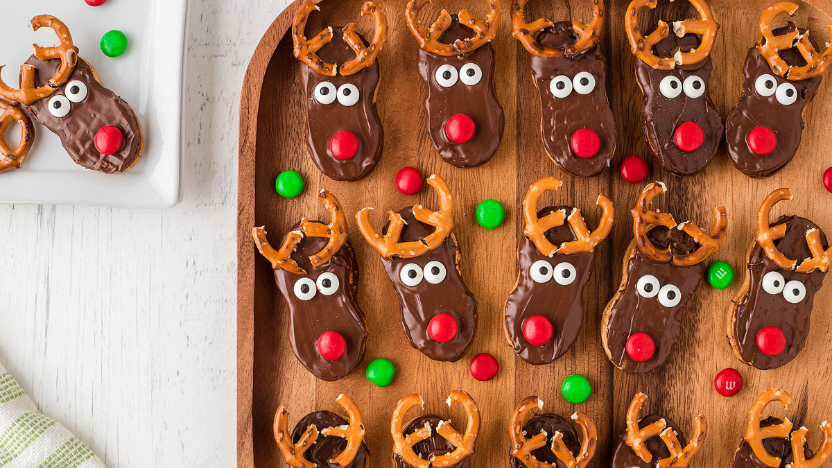 Chocolate-covered cookies decorated as reindeer with pretzel antlers, candy eyes, and red noses are arranged on a wooden tray with red and green candies.