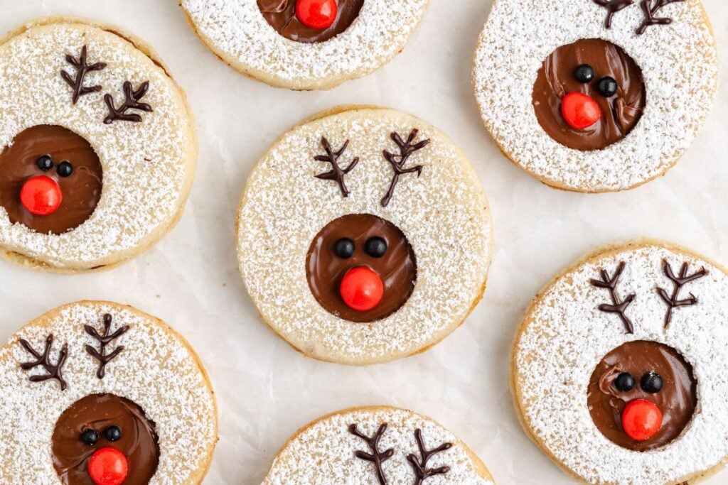 Round cookies decorated with powdered sugar, chocolate filling, red candy noses, and chocolate antlers to resemble reindeer faces, arranged on a light surface.