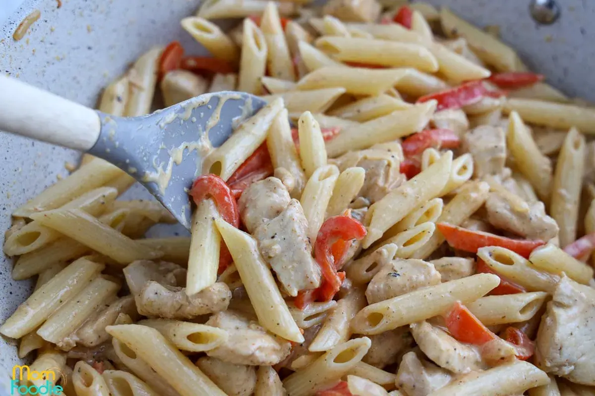 Penne pasta with pieces of chicken and red bell peppers in a creamy sauce being stirred with a spatula.